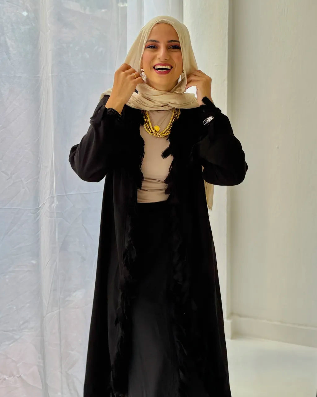 Woman wearing a black  set and beige hijab against a white curtain background