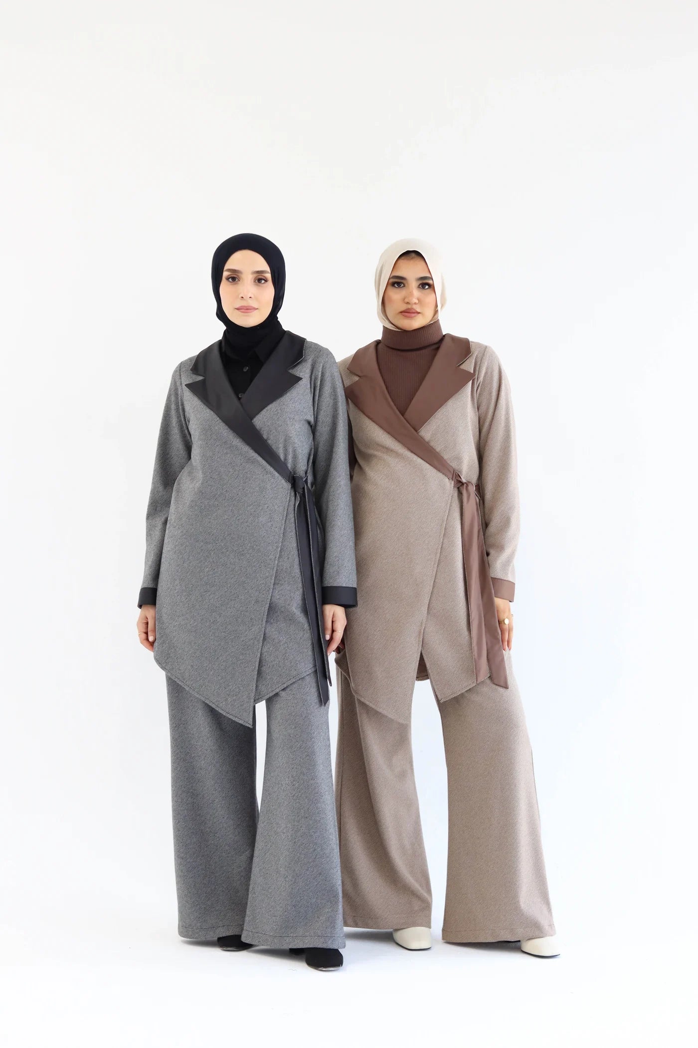 Two models wearing elegant two-piece wrap sets: one in medium grey and one in warm beige, both with asymmetric wrap tops, long sleeves, and wide-leg pants, full-length front view.